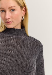 Blonde woman with blue eyes wearing a dark gray knitted turtleneck sweater and silver hoop earrings, neutral background.