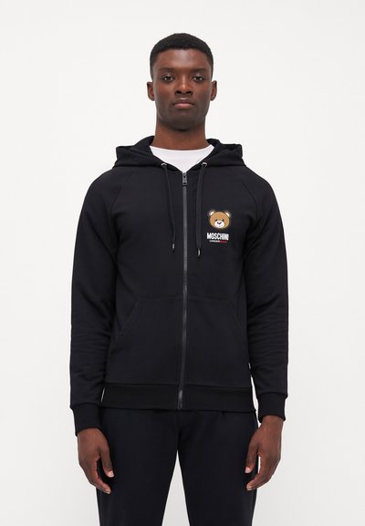 Černá mikina s kapucí na zip, která má logo medvěda a nápis „Moschino“. Vyrobena z měkké látky s žebrovanými manžetami a předními kapsami.