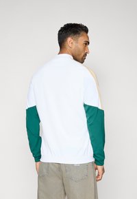 Verryttelytakki, jossa valkoinen runko, vihreät hihat ja kullanväriset yksityiskohdat. Urheilullinen muotoilu ja sileä pinta.