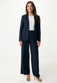 Marinblå skräddarsydd kostym med en figursydd kavaj och vida byxor, i kombination med en stickad vit topp och guldiga klackar med remmar. Slät textur, strukturerad design.