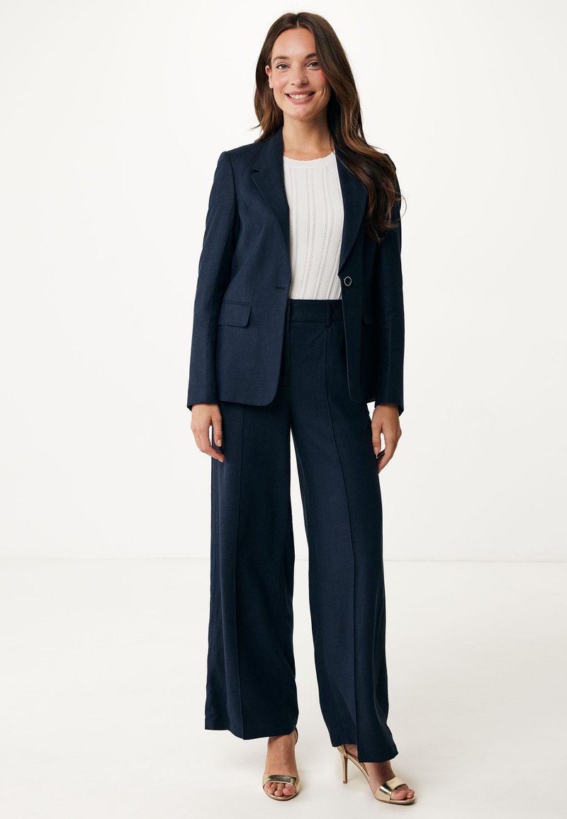 Marinblå skräddarsydd kostym med en figursydd kavaj och vida byxor, i kombination med en stickad vit topp och guldiga klackar med remmar. Slät textur, strukturerad design.