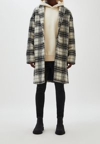 Persona vestida con una sudadera beige, un abrigo largo de cuadros gris y negro, jeans skinny negros y botas negras de tobillo sobre un fondo sencillo.