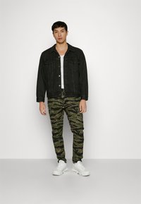 Jachetă din denim negru peste un top alb fără mâneci, asortată cu pantaloni verzi în stil camuflaj și adidași albi. Printre caracteristici se numără închiderile cu nasturi și buzunarele.