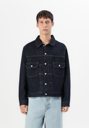 Joven de pie, vistiendo una chaqueta de mezclilla oscura con bolsillos en el pecho abotonados y jeans azul claro, contra un fondo blanco liso.