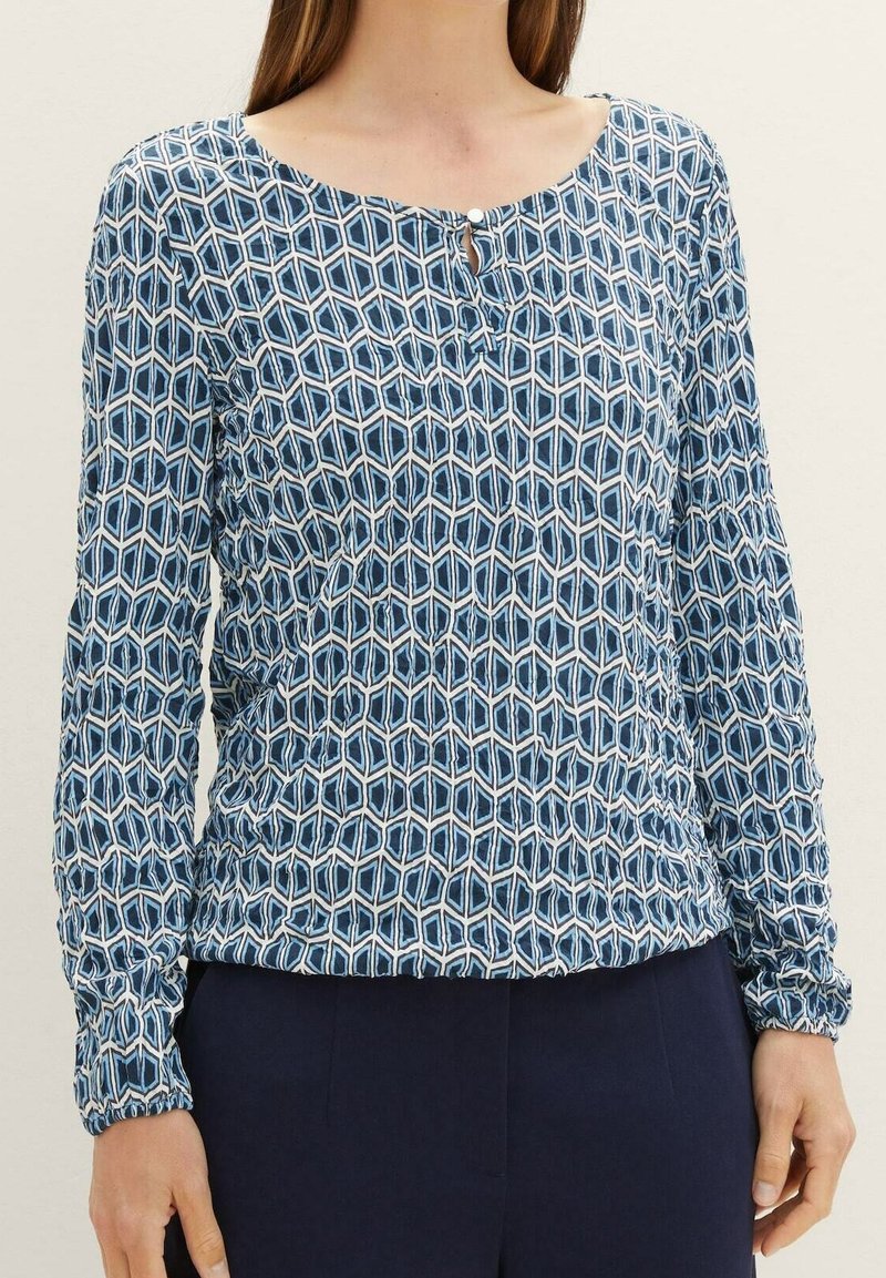 Haut à manches longues en tissu léger, avec un motif géométrique bleu, un décolleté en forme de scoop agrémenté d'un détail bouton, et des poignets froncés.