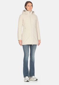 Giacca isolante beige chiaro con cappuccio, zip completa e due tasche laterali, abbinata a jeans a campana blu e scarpe da ginnastica grigie.