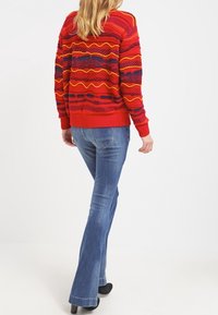 Červený pletený svetr s texturovanými vzory ve žluté a fialové barvě, doplněný modrými rozšířenými džínami. Černé podpatky dokončují vzhled.