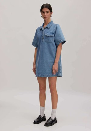 Vrouw draagt een blauw denimjurkje met korte mouwen, een kraag, een voorzak, witte sokken en zwarte loafers, staand op een effen lichte achtergrond.