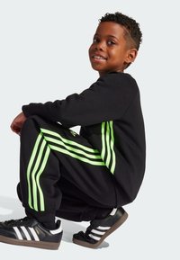 Schwarzes Sweatshirt und schwarze Jogginghose mit drei vertikalen neongrünen Streifen. Schwarze Schuhe mit weißen Streifen und strukturierten Sohlen. Kind in hockender Pose.