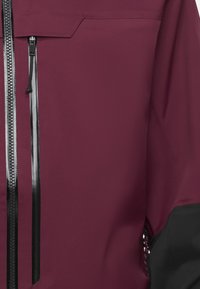 Bordeauxfarbene Jacke mit einem eleganten, matten Finish. Verfügt über einen auffälligen Reißverschluss mit schwarzer Einfassung und einer schrägen Brusttasche. Glatte Textur.