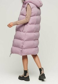 Lång, ärmlös, ljuslila pufferväst med huva, med en quiltad design och sidofickor med dragkedja. Buren med en åtsittande brun topp och ankelskor.