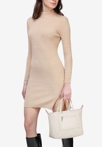 Beige rippet kjole med høj hals, lange ærmer og sideslids. Modellen holder en cremet håndtaske med en lynlås foran. Knæhøje sorte støvler.