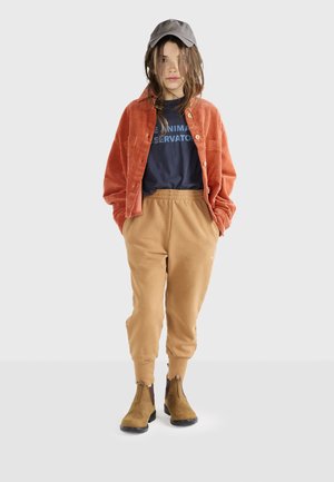 Orange kordflorskjorta med knappar, marinblå grafisk t-shirt, beige joggerbyxor och bruna ankelskor, tillsammans med en grå keps.