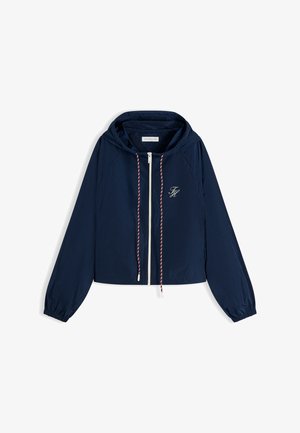 Chaqueta ligera azul marino con capucha, cremallera frontal, cordones rojos y blancos, puños elásticos y un pequeño logo blanco bordado en el pecho.