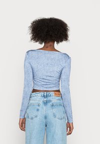 Top cropped azul de manga longa com um padrão texturizado, combinado com jeans claros de cintura alta que apresentam uma etiqueta castanha na parte de trás.