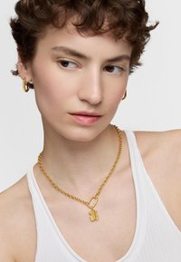 Collier en chaîne dorée avec un pendentif texturé au design géométrique, porté sur un haut sans manches blanc.