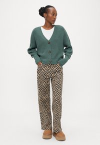 Πράσινη πλεκτή ζακέτα με κουμπιά, συνδυασμένη με καφέ ριγέ παντελόνι φαρδιάς γραμμής και ταμπά παπούτσια. Απλό λευκό κοντομάνικο μπλουζάκι από μέσα.