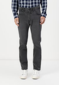 Mann iført mørkegrå jeans, hvite joggesko og en blå og hvit rutete skjorte, stående foran en hvit bakgrunn.