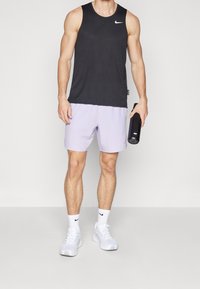 Débardeur de sport noir, short violet clair, baskets blanches, tenant une bouteille d'eau foncée. Logo Nike sur le débardeur et le short.