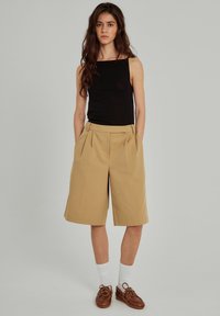 Tan veckade shorts med rak skärning, bärs med en åtsittande svart linne och bruna skor. Vita strumpor synliga, enfärgad bakgrund.