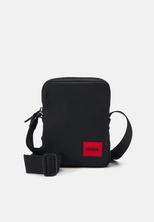 Black fabric crossbody bag with a rectangular shape, adjustable strap, and a red logo patch featuring the word "HUGO." Smooth texture.