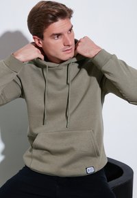 Khaki mikina s kapucňou a šnúrkami, klokanie vrecko a rebrované manžety; textúrovaná tkanina s jemným leskom. Na štítku je branding po boku.