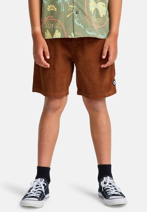 Braune Cordshorts aus Cord mit einer strukturierten Oberfläche, getragen mit schwarzen Sneakers und weißen Schnürsenkeln. Das Oberteil zeigt ein bunt gemustertes Hemd.