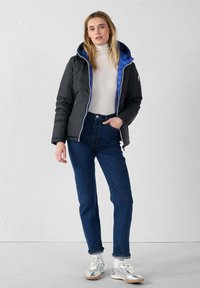 Giacca a vento nera con fodera blu, chiusura con zip e cappuccio. Abbinata a jeans blu a vita alta e scarpe da ginnastica argentate.