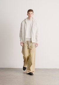 Laza öltözék világosszürke kapucnis pulóverrel, fehér bordázott pulóverrel és bézs bő szárú nadrággal. A modell fekete cipőt visel. Minimalista dizájn.