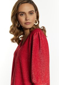 Rode glitter top met een V-hals en pofmouwen. Heeft een zachte textuur en glanst onder het licht, wat de levendige kleur versterkt.