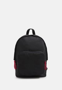 Sac à dos noir de forme arrondie, doté d'une poche avant en maille et de tirettes de fermeture éclair rouges. Fabriqué en matériau léger.