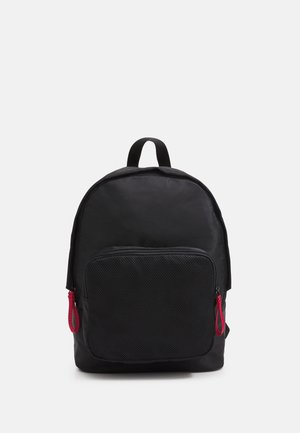 Sac à dos noir de forme arrondie, doté d'une poche avant en maille et de tirettes de fermeture éclair rouges. Fabriqué en matériau léger.