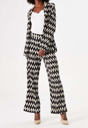 Vrouw die een blazer met zwart-wit zigzagpatroon draagt, wijde broekspijpen, een wit topje met kanten randje en beige hakken.