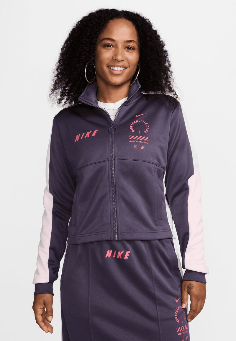 Chaqueta de pista Nike en púrpura oscuro con acentos en gris claro y rosa. Cuenta con cierre de cremallera, logo impreso y un diseño deportivo con bloques de color.