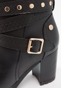 Bottine en cuir noir à talon haut avec des brides texturées, une boucle dorée et des détails en clous dorés.