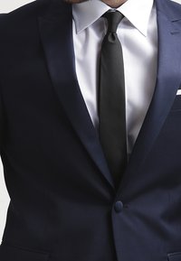 Granatowa marynarka, czarny krawat, biała koszula. Gładki materiał o strukturalnym wzornictwie, z jedynym zapięciem na guzik i klapami w kształcie notcha.