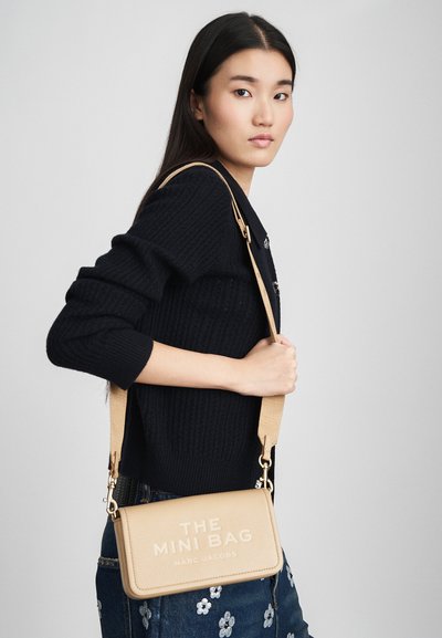 Młoda kobieta w czarnym swetrze z dzianiny i kwiecistych dżinsach, trzymająca beżową torebkę na ramię oznaczoną "THE MINI BAG MARC JACOBS".