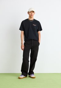 Tricou negru cu un grafic mic, pantaloni negri și adidași multicolori. Bărcuță deschisă, pandantiv din argint, stând pe un podea verde.