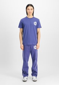 Vijolična bombažna majica s kratkimi rokavi in ujemajoči se trenirke z belim tiskom "ALPHA". Hlače imajo bele stranske obrobe, oba izdelka pa imata sproščen kroj.