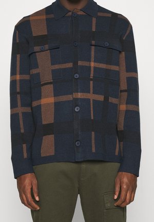 Granatowo-brązowa koszula w kratę zapinana na guziki z kieszeniami na piersi, noszona z oliwkowozielonymi spodniami na osobie stojącej na białym tle.