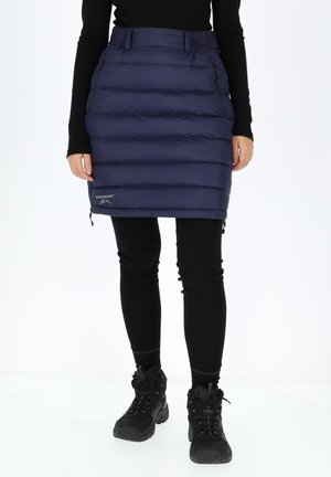 Navy puffer skirt with horizontal quilted design, zippered pockets, and adjustable waistband, paired with black leggings and sturdy boots.