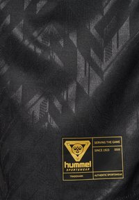 Crni sportski materijal s suptilnim geometrijskim uzorkom i zlatnom hummel logotip oznakom na kojoj piše „Serving the game since 1923“ i „Authentic sportswear.“