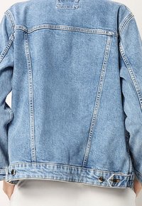 Vaaleansininen farkkutakki takaa nähtynä, jossa on pystysuoria ja vaakasuoria sauman yksityiskohtia sekä säädettävät napilla varustetut vyötäröntabit.