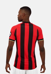 Maillot à rayures verticales rouges et noires avec des manches courtes, un col rond et des accents jaunes sur les bords des manches. Le matériau semble léger.