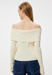 Cremfarbener, schulterfreier Rippstrickpullover mit langen Ärmeln, figurbetonter Silhouette, strukturiertem Stoff, kombiniert mit dunkler Jeans.
