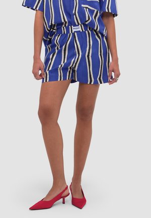 Osoba nosząca niebieskie, białe i czarne pionowo paskowane szorty oraz pasujący top, zestawione z czerwonymi, spiczastymi sandałkami na obcasie typu slingback.