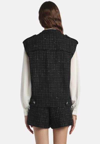 Veste en tweed noir avec un motif texturé, épaulettes aux épaules et accents de boutons métalliques, portée sur un chemisier de couleur claire et un short.