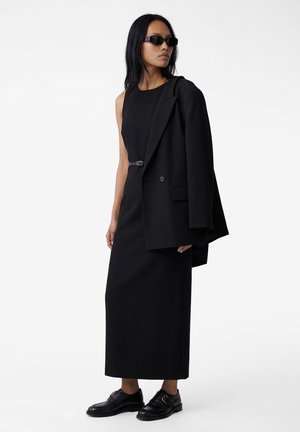 Vrouw draagt een lange zwarte mouwloze jurk, een zwart blazer over de schouder gehangen, zwarte zonnebril en zwarte leren schoenen.