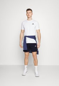 Balta Adidas t-kreklu ar mazu logotipu, tumši zila šortus un zilu džemperi, kas sasieti ap vidukli; pabeidz ar baltām sporta kurpēm un zeķēm.