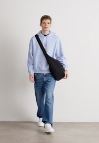 Čierna taška cez plece, vystužená textúra, nosená crossbody. Svetlomodrá mikina s kapucňou, šnúrkou a grafickým detailom. Voľné modré džínsy, biele tenisky.